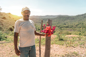 Medium shot of a Latino mature man with a mustache and a cap holding purple flowers in his hands in a mountainous area of Rivas Nicaragua - Powered by Adobe