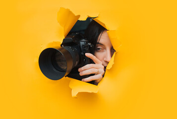 A young caucasian paparazzi girl holds a reflex camera and looks through a torn hole in yellow paper. The concept of embarrassment, shame for what he saw. Copy space. - obrazy, fototapety, plakaty