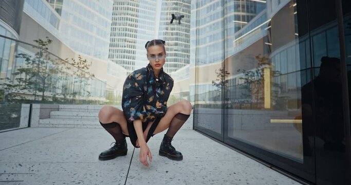 Urban style seductive girl wearing short skirt futuristic glasses posing in wind looking at camera standing and squats near high skyscrapers modern buildings. Attractive female fashion model