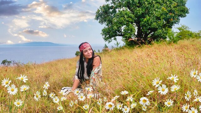 A young woman wearing a headscarf over long black hair and face glitter on her forehead, looks calm and relaxed while sitting in a field of long grass and daisies, with the ocean in the background.