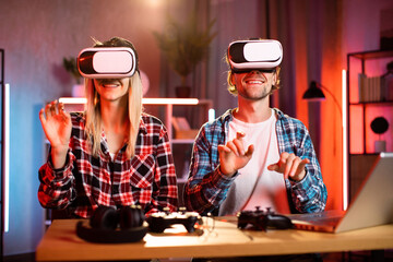 Young attractive couple playing games in virtual reality goggles. Handsome man and pretty woman sitting at table and touching virtual screen with fingers. Modern entertainment and fun at home.