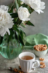 A bouquet of white peonies in a glass vase, a white ceramic mug with tea and pretzels