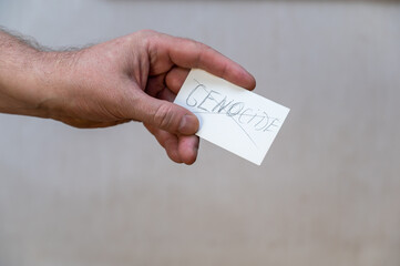 The word GENOCIDE is crossed out. A word written in jagged letters. A man's hand holds a white paper rectangle with text against a gray background