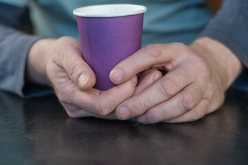 A grown man holds a lilac glass in his hands. A paper cup for hot drinks. Eco-friendly utensils concept. Man sitting at a black table indoors. Close-up. Selective focus.