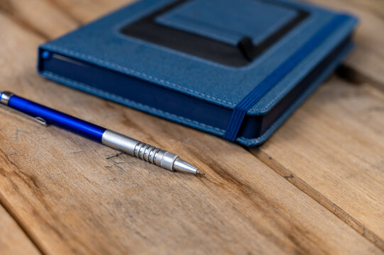 A blue diary and pencil against a wooden background. A closed hardcover notebook. A metallic silver-blue pencil rests on top of old cracked boards. Close-up. Selective focus.