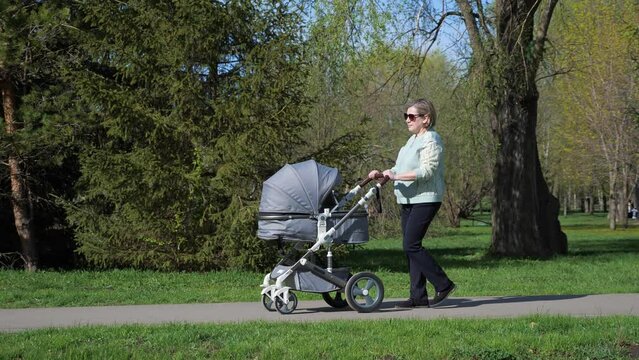 Grandma walks with stroller on road in city park with lush green trees in spring. Senior woman spends time with newborn granddaughter slow motion