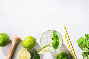 Flat-lay of mojito cocktail with lime, fresh mint and ice on white texture background. Summer refreshment citrus drink. Top view and copy space.