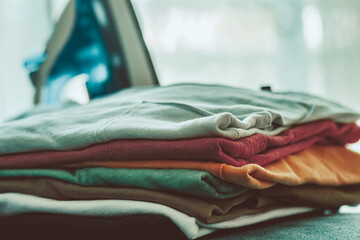 Closeup of folded colorful soft textile clothes placed on surface with electric pressing iron in bright bedroom at home on blurred background