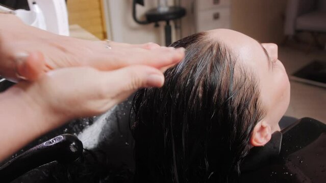 Woman hairdresser applies balm to black strands of lady client after washing hair in spa salon closeup. Professional step-by-step hair and scalp care