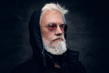 Photo of senior man hipster dressed in sunglasses and jacket with hood against dark background.