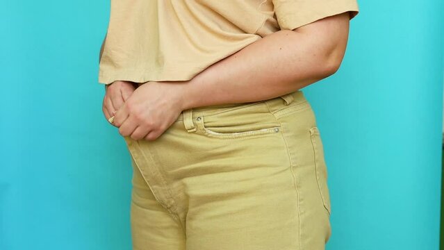 Side view of unrecognizable fat overweight woman wearing beige T-shirt, underwear, putting on, buttoning up jeans on blue background. Body positive, massage, obesity, weight loss, liposuction, diet.