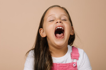Little girl with fully opened mouth and closed eyes yawning looking tired wearing bright pink jumpsuit and white t-shirt on beige background.