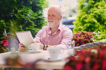 Shot of bearded active grandfather with tablet drinking tea sitting at table outdoors.