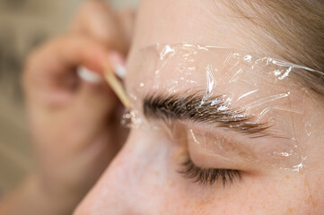The master applies the composition for laminating the eyebrows to the client with a wooden spatula. 
