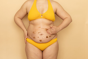 Fat woman in yellow swimsuit apply brown body cream on thick sagging stomach, beige background....