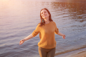 Happy woman relaxes with her arms out to sides on shore lake at sunset in rays of the setting sun. Enjoys the day and good mood in casual clothes laughing merrily