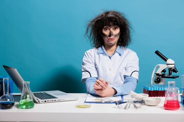Fototapeta premium Crazy mad biochemistry expert with messy hair and dirty face sitting at lab desk after experiment explosion. Maniac goofy looking chemist looking dumb while looking at camera on blue background.
