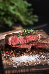 Medium-rare sliced beef steak with salt and herbs on a wooden board on a dark background.