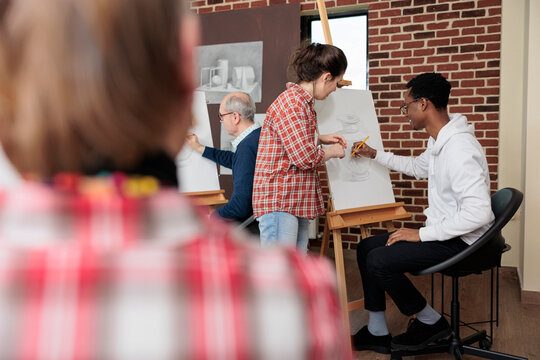 Artist teacher explaining drawing process to young student during creativity school course. Diverse team sketching vase model on white canvas using illustration technique. Creative concept
