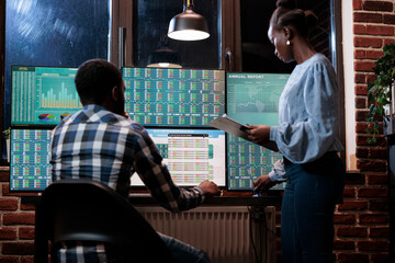 Investment company professional agents sitting at multi monitor workstation in office while analyzing financial data. African american coworkers discussing about possible stock selling.