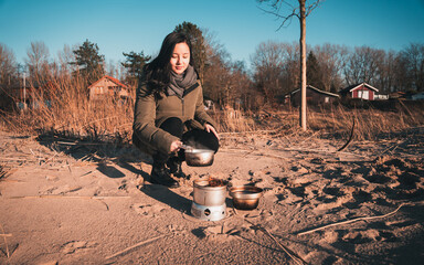 Young asian woman with warm clothes on is cooking with outdoor cooker on Harriersand beach at a riverbank during golden hour. Weser river blurred in the background. 