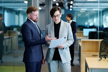 Young businessman pointing at business contract and discussing it together with businesswoman while they standing at office