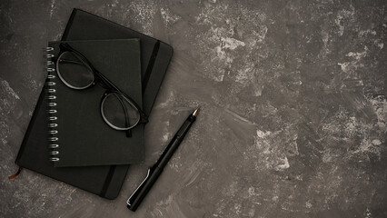 Stylish black spiral notebook, glasses, pen and copy space on grey cement background.