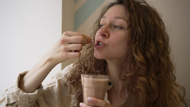 Curly woman takes greenery out of smoothie glass and eats. Young woman with blue eyes smells smoothie and cheerfully smiles at camera