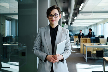 Fototapeta premium Portrait of beautiful young woman with short hair in formal suit holding digital tablet and looking at camera standing at office