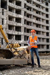a great worker in uniform and helmet stands by an excavator on a construction site.