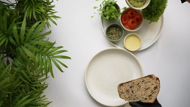 Hands in black rubber gloves put green guacamole on slice of rye whole grain bread with wooden spoon. Cooking ingredients placed on white tray top view