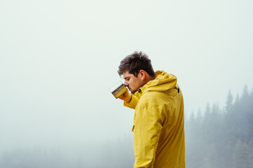 Male hiker in a yellow jacket drinks from a cup of hot tea on a background of misty view in the mountains.