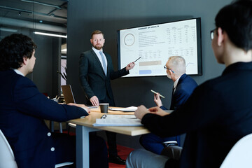 Young businessman in suit pointing at display screen with charts and performing with his presentation for colleagues at meeting room