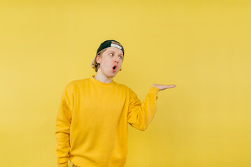 Shocked young man in a cap and yellow sweatshirt holds a copy space in his hand and looks away with a surprised face.