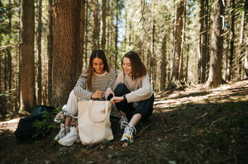 Two happy women in casual clothes sit in the woods and rest, look in the eco bag and rest