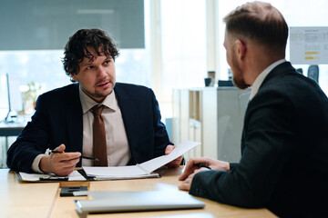 Young businessman in suit examining contract and discussing it with his partner at table at meeting