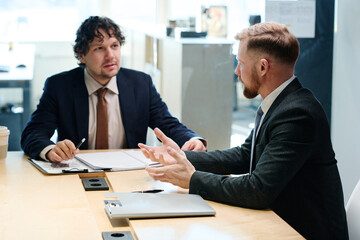 Two colleagues in formal suits discussing business plan in team at table during meeting at office