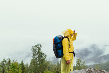 Man in a raincoat with a backpack on his back stands on a rock and looks away at the beautiful views and looks away.