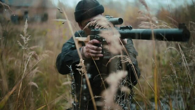 Army sniper sitting and aiming in grass carrying his rifle.