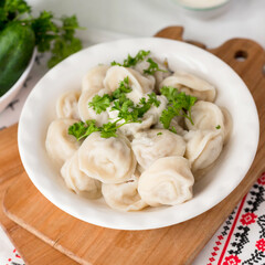 Dumplings in a clay plate on a wooden board on an embroidered tablecloth with vegetables (tomatoes, cucumbers, parsley) and sour cream with a vintage spoon