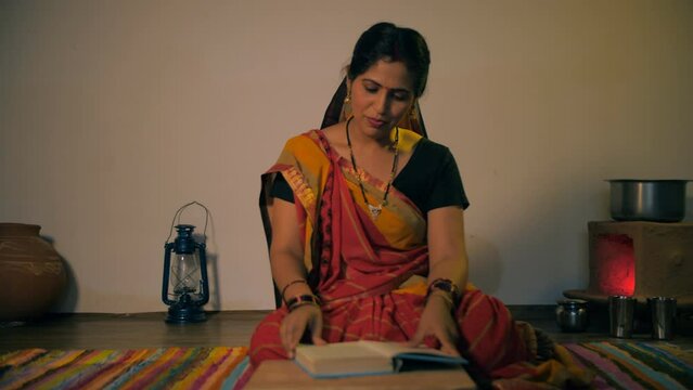 Confident Indian woman fluently reading a book sitting inside her kitchen. Medium shot of a young rural housewife in a saree reading Ramayan book - literate village woman