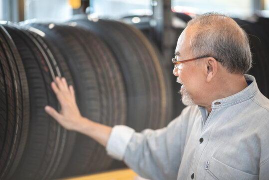 Smiling Senior Man With Beard And Spectacles Checking Car Tires From Rack In Garage While Getting Assistance From Young Male Mechanic