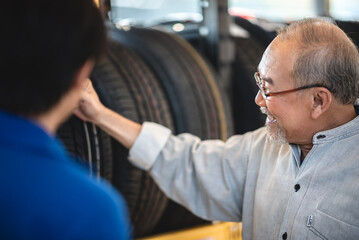 Obraz premium Smiling senior man with beard and spectacles checking car tires from rack in garage while getting assistance from young male mechanic