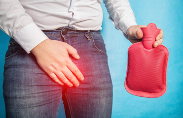 A man in a white shirt holds his groin and holds a red heating pad with hot water in his hand. The concept of treatment of male diseases in the genital area. Close-up - obrazy, fototapety, plakaty