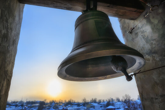 Heavy cast bell in the church bell tower against the yellow-blue sunset sky in the window opening. Close-up.