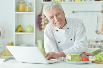 Elderly male chef cooking at table with laptop