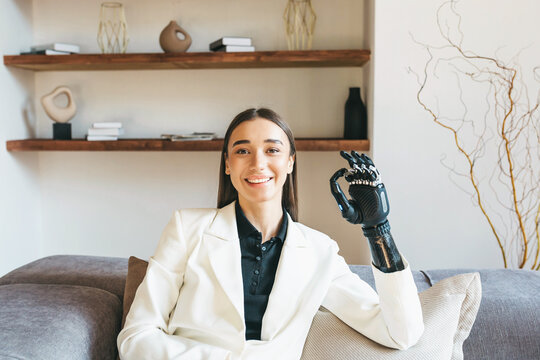 Portrait of a girl with disabilities. A beautiful young European girl is sitting on the sofa in casual clothes and shows the "OK" sign with a prosthetic bionic arm