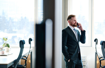 Young bearded manager in formal wear smiling while having conversation on mobile phone at office