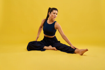Obraz premium Slender girl in preparing to take one leg forward fold pose of yoga, stretching on floor. Front view of brunette female sitting in asana of yoga, isolated on orange studio background. Concept of yoga.