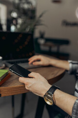 Businessman with crop hands messaging on smartphone with black screen while working remotely in coffee shop and browsing laptop 